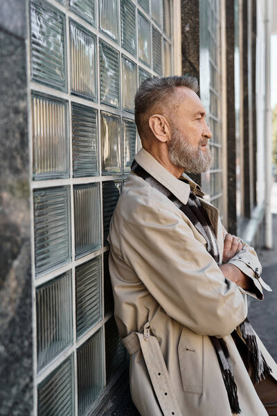 A fashionable senior man stands confidently against a glass brick wall in a vibrant city.