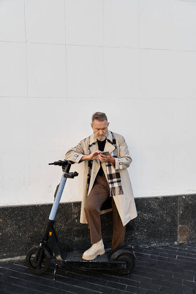 Старший мужчина в шикарном плаще сидит на электрическом скутере, поглощённом телефоном.