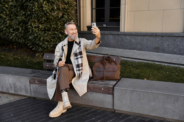 A dapper senior man in trendy clothing sits on a bench, engaging in a cheerful selfie.