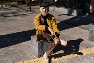 Güler yüzlü bir son sınıf öğrencisi kentteki taştan bir bankta rahat eder, modaya uygun giyinir..