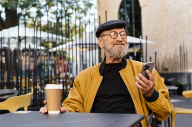 Son sınıf öğrencisi, modaya uygun ceketli ve şapkalı, kafe masasında oturuyor, kahvesini yudumluyor ve telefonla konuşuyor..