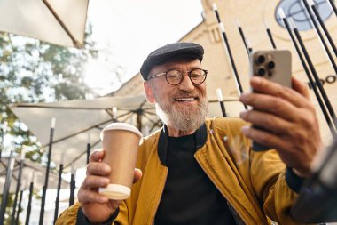 Üst sınıftan bir adam şık bir açık hava kafede oturmuş telefonuyla selfie çekerken geniş bir gülümseme sergiliyor..