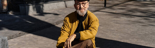 В модном ансамбле пожилой человек излучает очарование, сидя на каменной скамейке.