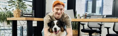 Genç kızıl saçlı bir adam modern ofis ortamında bir köpekle oynarken gülümsüyor..