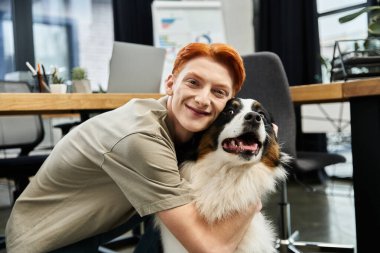 Kızıl saçlı genç bir adam modern bir ofiste çalışırken köpeğiyle oynar..