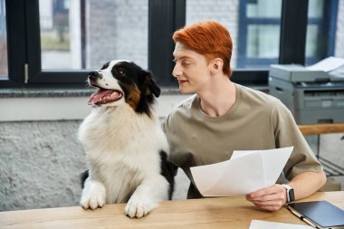 Kızıl saçlı genç bir adam modern bir ofiste köpeğiyle birlikte masasında oturuyor..