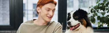 Kızıl saçlı genç yakışıklı adam masasında çalışırken neşeli bir köpekle bir anı paylaşıyor..
