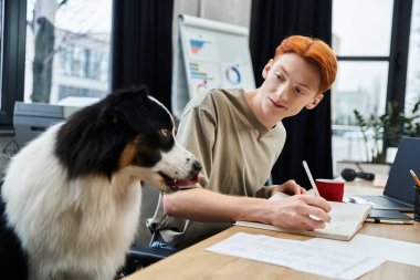 Kızıl saçlı genç bir adam bir ofise notlar yazıyor, köpeği yanında oturuyor..