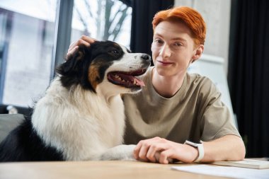 Genç kızıl saçlı bir adam ofis masasında bir köpekle neşeyle etkileşime geçiyor..