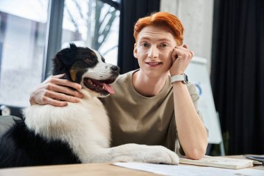 İşyerinde köpeğiyle vakit geçirirken kızıl saçlı genç bir adam gülümsüyor..