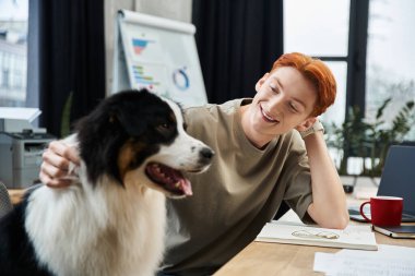 Genç kızıl saçlı bir adam modern bir ofiste neşeli bir köpeğe gülümsüyor..