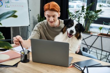 Kızıl saçlı genç adam iş yerinde yanında köpeğiyle gülümsüyor..