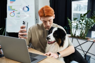 Kızıl saçlı, kendini işine adamış genç bir adam ofiste yoğun bir iş günü boyunca köpeğiyle etkileşim halindedir..