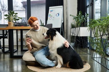Kızıl saçlı genç bir adam modern bir ofiste köpeğiyle uğraşırken yerde oturuyor..