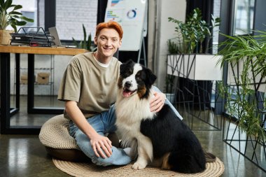Modern bir ofiste genç bir adam yerde oturuyor, köpeği ve bitkileriyle gülümsüyor..