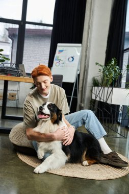 Kızıl saçlı genç bir adam neşeli bir ofiste köpeğiyle oynayarak mutlu bir şekilde çalışıyor..