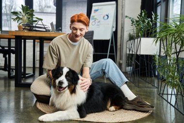Modern bir ofiste, kızıl saçlı genç bir adam köpeğiyle gülümser ve sıcaklığı arttırır..