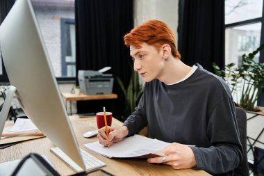 Dedicated young professional with red hair engrossed in important tasks at his office desk. clipart