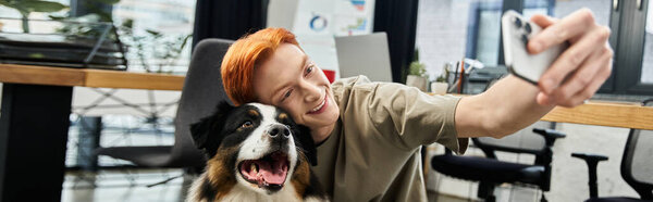 A young man with red hair smiles for a selfie with a joyful dog at work.