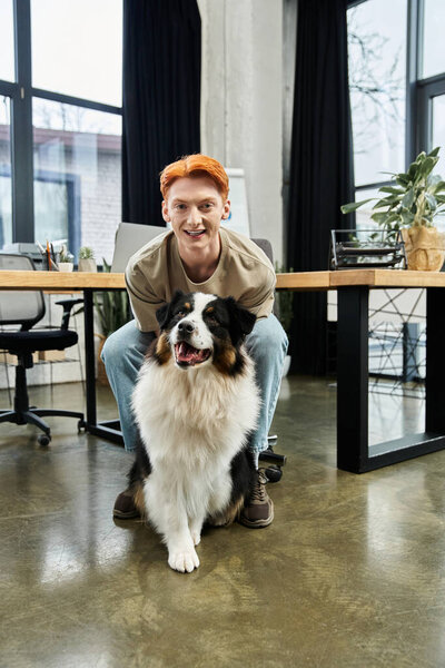 A young man with red hair enjoys time with his dog in a bright office space.