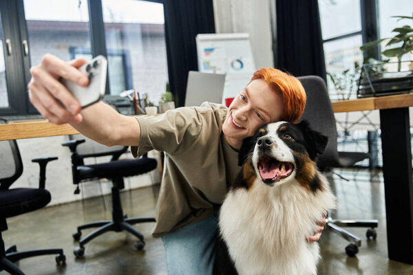 A young man with red hair captures a happy selfie with a friendly dog in a stylish office.