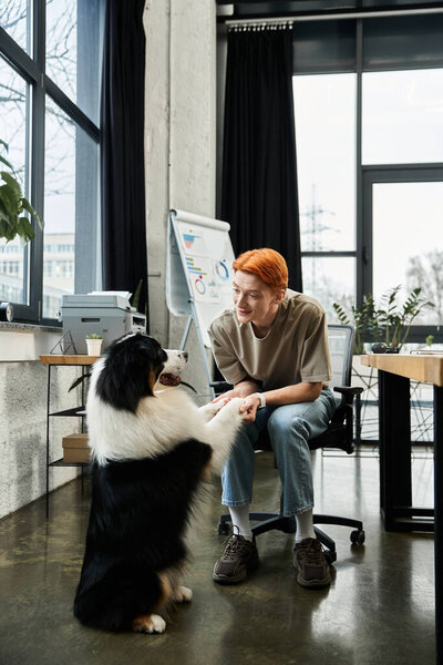 A dedicated young man with red hair enjoys a playful moment with a dog in an office.