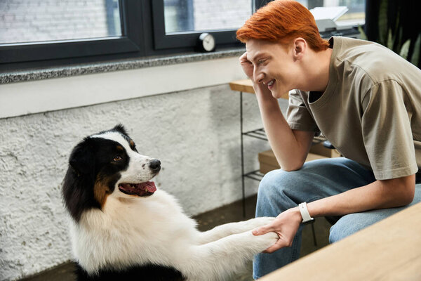A young man with striking red hair enjoys a playful moment with a friendly dog while at work.