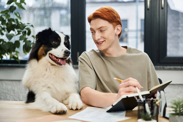 A handsome young man with red hair writes in a notebook while his dog watches closely.