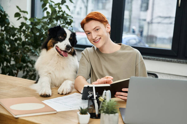 A young red haired man smiles at his playful dog as he completes his tasks in a sleek office.