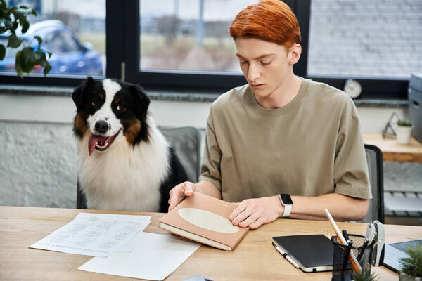 A young man with red hair works at his desk with a friendly dog by his side.