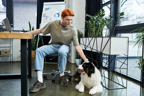 Молодой человек с рыжими волосами наслаждается своей работой в современном офисе, гладя собаку..