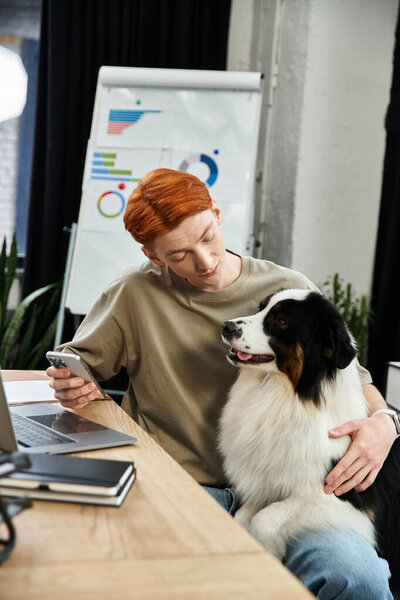 In a bustling office setting, a young red haired man engages with his dog while attending to tasks.
