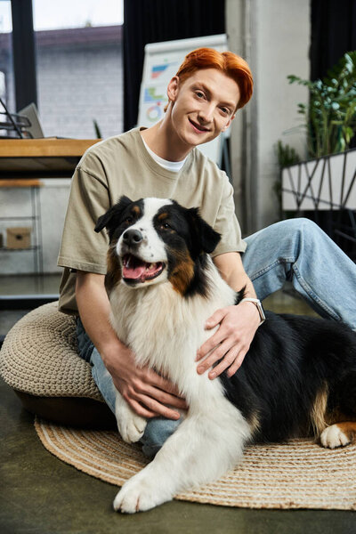 Red haired man sits on the floor, smiling and playing with a friendly dog in a bright office.