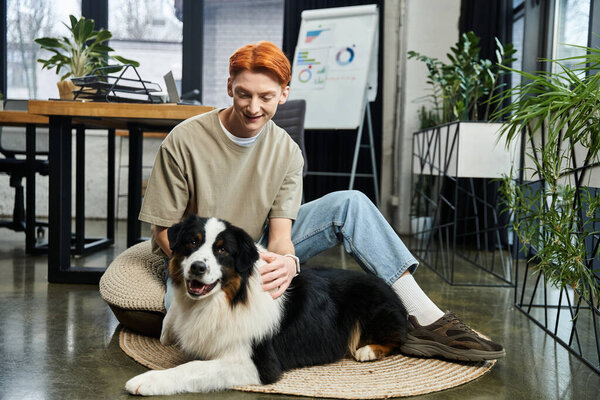 In a modern office, a young man with red hair shares smiles with his dog, fostering warmth.
