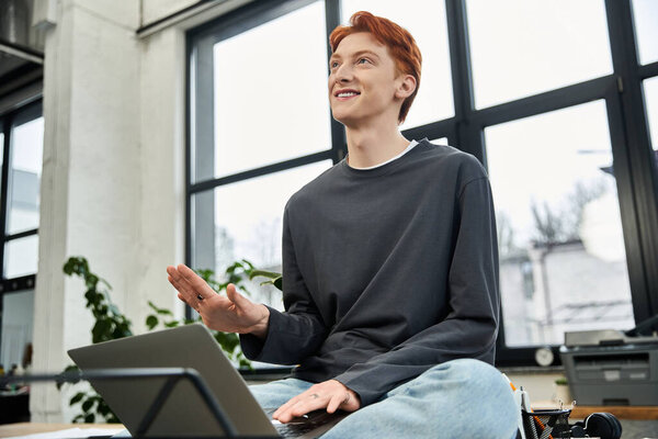 Молодой рыжий мужчина с энтузиазмом сидит за своим столом в светлом офисе.