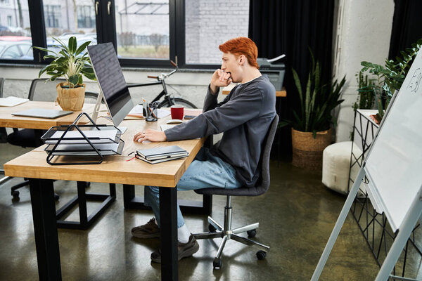 In a modern office, a focused young man with red hair works diligently at his desk.