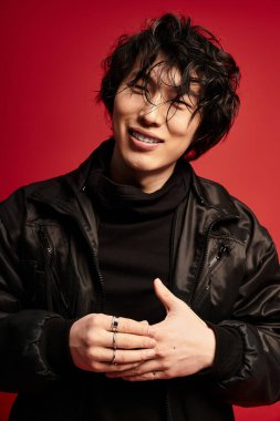 Siyah kıyafetli genç bir Asyalı stüdyoda poz veriyor. Kırmızı arka planda, kurnazca gülümsüyor..
