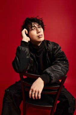 Şık genç adam kırmızı bir arka plana karşı bir sandalyede dinleniyor, kendine güveni ve modern zarafeti yayıyor..