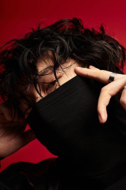 Koyu renk saçlı Asyalı adam yüzünü kırmızı bir stüdyoda kısmen kapatmak için siyah bir balıkçı yaka giyiyor.