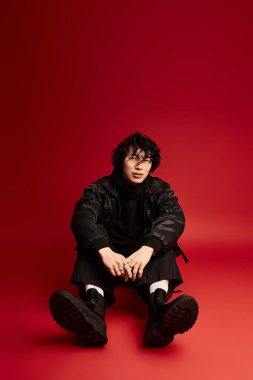 Şık siyah kıyafetli Asyalı genç adam kırmızı stüdyo arkaplanında oturur, serinkanlı ve kendine güvenir..