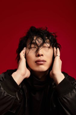 Gözü kapalı şık genç bir adam, koyu kırmızı bir arka planda şakaklarına dokunur. Stüdyo çekimi.