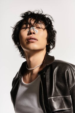 Burnu alçılı şık Asyalı bir adam stüdyoda poz veriyor. Deri ceket ve şık aksesuarlar giyiyor..