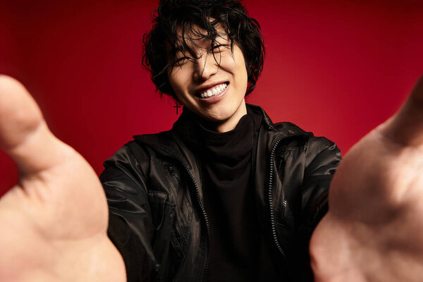 A stylish young asian man smiles widely while reaching out with both hands in a studio.