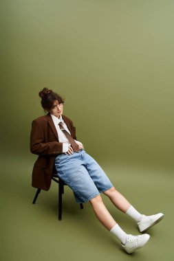 Moda kıyafetleri içinde genç bir kadın zarif bir şekilde sandalyede poz veriyor, kendine güveni ve tarzı var..