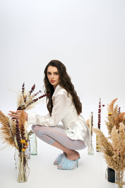 Молодая женщина с каштановыми волосами изящно приседает среди элегантных цветов, вызывая очарование весны..