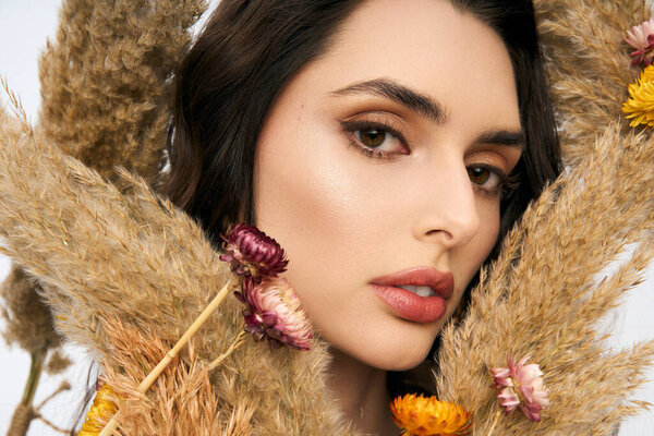 Молодая женщина с каштановыми волосами грациозно стоит среди цветущих цветов, демонстрируя весеннюю красоту.