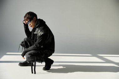 Yakışıklı Asyalı adam modern bir stüdyoya çömelmiş, yumuşak ışıklandırma altında şık bir kıyafet sergiliyor..