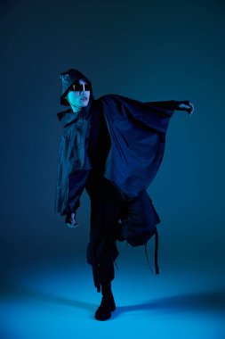 Yakışıklı Asyalı genç adam mavi ışıklarla aydınlatılmış çarpıcı siyah bir elbiseyle dramatik bir şekilde poz veriyor..