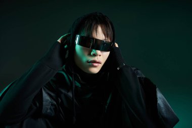 Genç Asyalı adam çarpıcı güneş gözlüğü ve siyah bir kıyafet giyerken modern ve gergin bir görünüm sergiliyor..