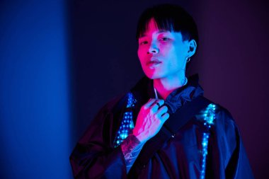 Kendine güvenen genç Asyalı adam, eşsiz moda anlayışını sergileyerek renkli bir arkaplana karşı duruyor..